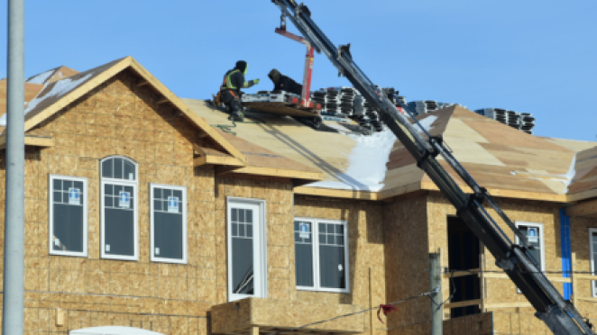 Construction crew working on the roof of a new residential building, with a long crane arm extending up to deliver materials.