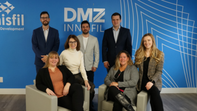 Group of seven professionals posing in an office setting in front of a blue wall with “DMZ Innisfil” branding, with four standing and three seated on chairs.