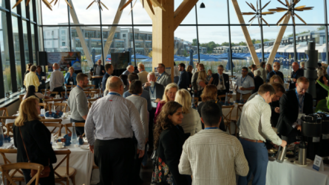 A large group of people networking at an indoor event with floor-to-ceiling windows, round tables, and a waterfront view in the background